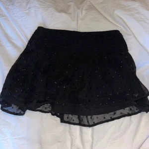 Svart volangkjol - Svart minikjol med små glitterprickar och volangdetaljer. Kjolen har en elastisk midja för bekväm passform. Oanvänd med lappar kvar 