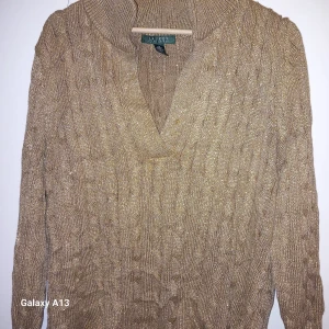 Beige stickad tröja från Ralph Lauren - Säljer en snygg beige stickad tröja från Ralph Lauren med ett subtilt glittrigt inslag. Tröjan har en v-ringad design och långa ärmar, perfekt för kyligare dagar. Den är tillverkad i en blandning av bomull och polyester med metalliska detaljer.