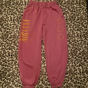 Rosa mjukisbyxor med Nirvana-tryck - Säljer rosa mjukisbyxor med elastisk midja. Byxorna har ett gult Nirvana-tryck längs benet och tre smiley-ansikten. Perfekta för en avslappnad stil. Har små fläckar vid låren.