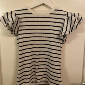 Randig topp från Holly & Whyte - Säljer en snygg vit och svart randig topp från Holly & Whyte by Lindex. Toppen har korta volangärmar och är ribbad för en skön passform. Perfekt för en stilren look!