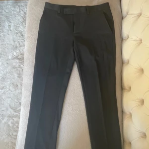 Svarta kostymbyxor från Dressmann - Snygga svarta kostymbyxor från Dressmann i slim fit. Byxorna har en klassisk design med knappar bak och en stilren look. Perfekta för en dressad stil.