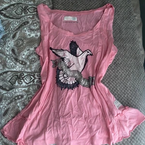 Rosa tanktopp från Odd Molly - Säljer en söt rosa tanktopp från Odd Molly med ett tryck av en fågel och texten 'In Dodo We Trust'. Toppen har en avslappnad passform och är perfekt för en ledig stil. Materialet är mjukt och bekvämt.