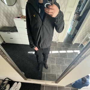 Svart vindjacka från Lyle & Scott - Säljer en stilren svart vindjacka från Lyle & Scott med dragkedja och huva. Jackan har ett gult broderat emblem på bröstet och är perfekt för blåsiga dagar. Den har långa ärmar och elastiska muddar för en bekväm passform. Jackan är storlek S fast jag  hade storlek XS och passade perfekt!👍🏻
