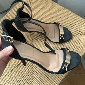 Svarta klackskor med gulddetaljer - Eleganta svarta sandaler med gulddetaljer och öppen tå. De har en smal rem runt ankeln och en dekorativ guldfärgad kedja framtill. Använda 1 gång, storlek 37🤗