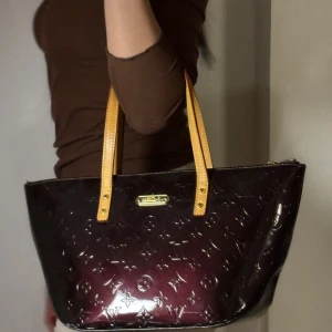 Mörklila handväska med monogram - Snygg mörklila handväska med glansigt monogrammönster och kontrasterande gula handtag. Perfekt för att ge din outfit en lyxig touch. Väskan har en dragkedja för säker förvaring.