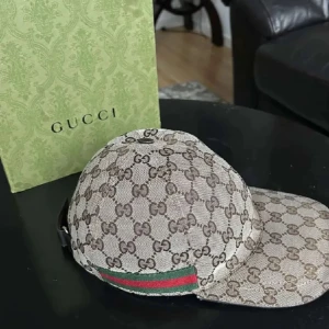 Beige keps från Gucci - Snygg beige keps från Gucci med det ikoniska GG-mönstret. Kepsen har en justerbar rem baktill i skinn och en grön och röd randdetalj på sidan. Perfekt för att ge din outfit en lyxig touch.