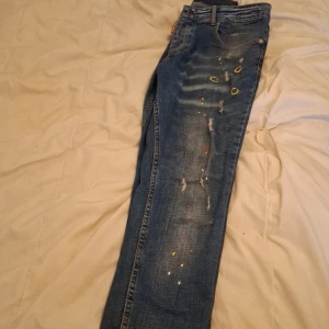 Dsquared2 jeans - Säljer ett par coola blå jeans med slitningar och färgstänk i olika färger. Jeansen har en straight passform och är dekorerade med små hål och slitna detaljer för en edgy look. Perfekta för dig som vill ha en unik stil.