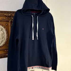 Mörkblå hoodie från Tommy Hilfiger - Säljer en snygg mörkblå hoodie från Tommy Hilfiger med vit dragsko och logotypdetalj på bröstet. Nederkanten har ett band med Tommy Hilfiger-loggan i rött, vitt och blått. Perfekt för en avslappnad stil. Helt ny, pris lapp på, nypris 70 euro. 