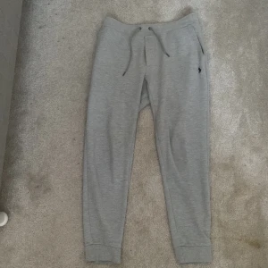 Grå mjukisbyxor från Ralph Lauren - Snygga grå mjukisbyxor från Ralph Lauren med dragsko i midjan och en liten broderad logga på sidan. Byxorna har en avslappnad passform och är perfekta för en bekväm stil. De har också fickor på sidorna för extra funktionalitet.