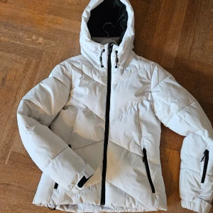 Vit jacka från McKinley strl 36  - Säljer en snygg vit jacka från McKinley med dragkedja och huva. Jackan har en quiltad design och praktiska fickor med dragkedjor. Perfekt som skidjacka med snölås och liftkortsficka.