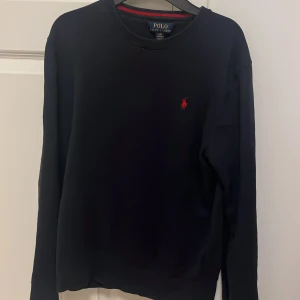 Mörk blå tröja från Ralph Lauren - Säljer en stilren svart tröja från Ralph Lauren med det klassiska röda broderade logotypen på bröstet. Tröjan har långa ärmar och en rund halsringning. Perfekt för en avslappnad och snygg look. L i barn storlek men är som S-XS