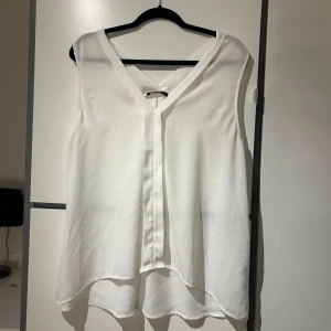 Vit ärmlös blus - Säljer en stilren vit ärmlös blus med v-ringning. Blusen har en luftig passform och är tillverkad i ett lätt material.