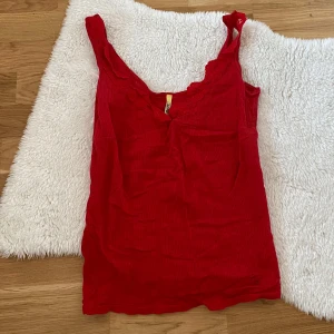 Röd ribbad linnetopp med spetsdetaljer - Snygg röd ribbad linnetopp med spetsdetaljer längs halsringningen. Perfekt för en somrig look eller som ett lager under en kofta. Toppen har en v-ringad design och ärmlös stil. 