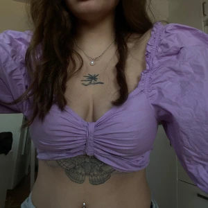 Lila kortärmad topp från Monki - Säljer en söt lila topp från Monki med puffiga korta ärmar och knytning framtill. Toppen är croppad och har en smickrande passform. Perfekt för en trendig look!