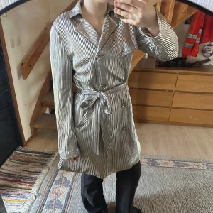 Randig trenchcoat med knytband - Snygg randig trenchcoat i silver och svart med knytband i midjan. Jackan har långa ärmar och knappar framtill. Perfekt för en stilren look.