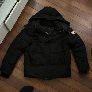 Svart dunjacka från Canada Goose - Säljer en svart dunjacka från Canada Goose med huva och dragkedja. Jackan har en klassisk design med långärmade ärmar och en logotyp på ärmen. Perfekt för kalla dagar. Obs javkan saknar en knapp men det syns knappt den går att stänga som vanligt 