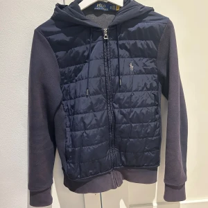 Mörkblå jacka med huva från Polo Ralph Lauren - Snygg mörkblå jacka med huva från Polo Ralph Lauren. Tröjan är i xs och passar till allt, perfekt nu till våren. Cardiganen är lite solbränd som det syns på bilder. Sen har den även 2 defekter på bild 4 och 5 men dem syns inte när man använder den, annars är det bra skick. Pris går att diskutera vid snabb affär.