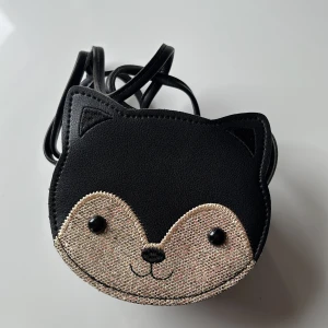 Svart axelväska i form av en katt för barn - Svart axelväska i form av en katt! Väskan har ett glittrigt ansikte med söta detaljer som små svarta knappar till ögon och en dragkedja med ett hjärtformat hänge. Perfekt för barn 