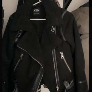 Svart jacka från Zara - Säljer en snygg svart jacka från Zara med fuskpälsfoder och dragkedjedetaljer. Jackan har en cool bikerstil med bälte i midjan och långa ärmar. Perfekt för höst och vinter när du vill hålla dig varm och stilren. Passar både till vardags och fest!