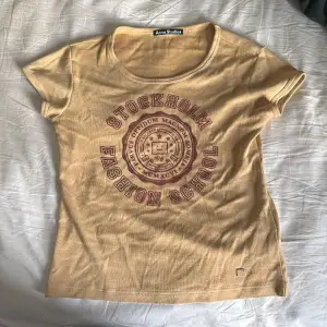 Säljer en snygg beige t-shirt från Acne Studios med tryck 'Stockholm Fashion School' i rött på framsidan. Den är i bomull och har en normal passform med korta ärmar. Perfekt för en avslappnad stil!