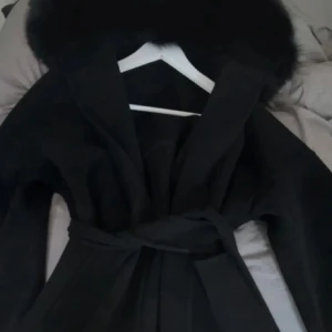 Svart kappa med pälskrage - Säljer en svart kappa med en äkta pälskrage. Den har ett bälte i midjan för en snygg passform och är perfekt för kyliga höst- och vinterdagar. Kappan är normal i storlek. Fint skick inga defekt, samt rök och djurfri.
