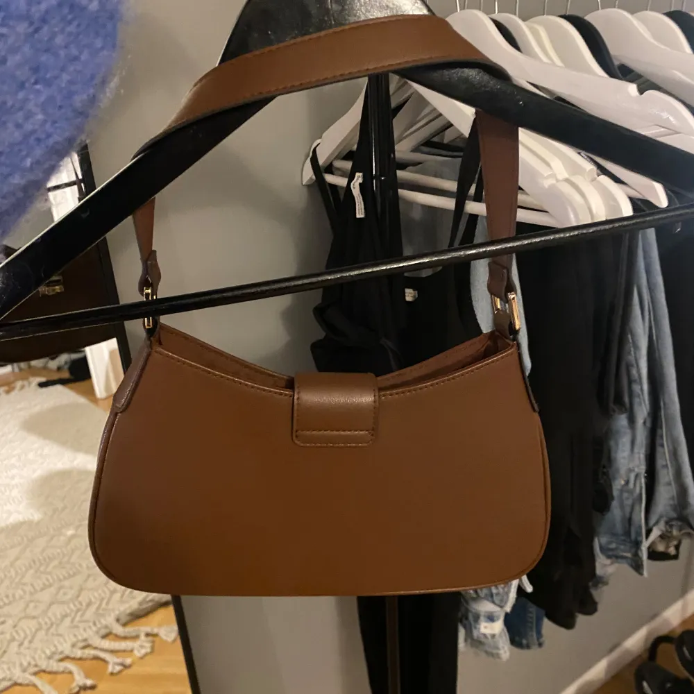 Säljer en snygg brun axelväska i skinn med gulddetaljer. Väskan har en stilren design med ett spänne framtill. Perfekt för både vardag och fest! Använd fåtal gånger!. Laukut & Käsilaukut.