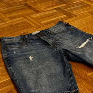 Blå jeansshorts med slitningar - Säljer ett par snygga blå jeansshorts från Jack & Jones. De har coola slitningar på framsidan som ger en avslappnad utseende. Perfekta för sommaren och vårens varma dagar. Shortsen har en klassisk femficksdesign och är i mycket bra skick. Regular Fit i storlek M!