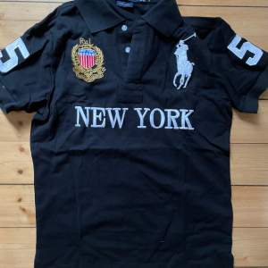 Säljer en snygg svart pikétröja från Polo Ralph Lauren. Den har ett stort nummer 5 på ärmen och texten 'NEW YORK' på framsidan. Tröjan är broderad med ett emblem i guld och rött. Perfekt för en stilren och sportig look!