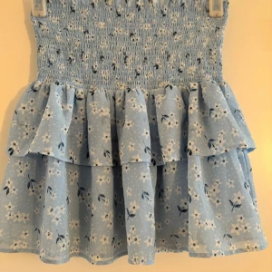 Blommig blå kjol med volanger - Superfin blå kjol med blommigt mönster och volanger. Den är i storlek 158/164 och gjord av 100% polyester. Perfekt för sommardagar eller en söt vårlook. Den har en smockad midja som ger en bekväm passform. 🌸Köparen betalar frakt. 