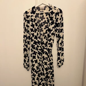 Mönstrad klänning från Fabienne Chapot - Säljer en superfin långklänning från Fabienne Chapot i storlek 36. Klänningen är i 100% viskos och har ett coolt svart blommönster på en beige bakgrund. Den har långa ärmar och en omlott-design som ger en snygg passform. Perfekt för både vardag och fest! Helt ny och aldrig använd! Nypris 1900kr