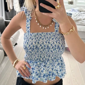 Blommig topp med volangdetaljer - Säljer en superfin blommig topp i blått och vitt. Den har smockad passform och volangdetaljer vid axelbanden och nederkanten. Perfekt för sommardagar eller en avslappnad vårlook. Toppen är ärmlös och har en söt, feminin stil.