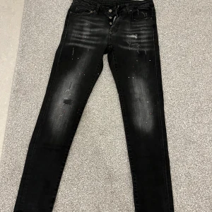 Svarta jeans från Dsquared2 - Säljer ett par svarta jeans Dsquared2 liknanade i bra skick. De har en cool, sliten look med ljusare partier och små färgstänk. Jeansen har en slim passform och är perfekta för både vardag och fest. De har knappar i gylfen och klassiska fem fickor.