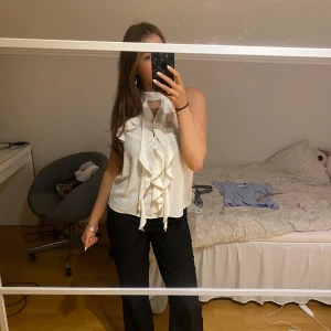 Vit blus med volangdetaljer - Säljer en elegant vit blus med volangdetaljer framtill. Den är ärmlös och perfekt för både fest och vardag. Blusen har en lös passform och är gjord i ett lätt och bekvämt material. Använd enstaka gånger. 