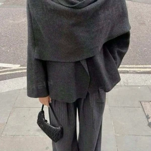 Mörkgrå kort kappa - Säljer en elegant mörkgrå kappa i ull. Den har en unik design med en draperad framsida (halsduk som sitter på kappans ena arm) och långa ärmar. Perfekt för höst och vinter, och ger en stilren look. Passar både till vardags och festliga tillfällen. Passar XS-M