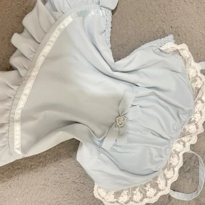 Ljusblå klänning med volanger och hjärtformad detalj - Söt ljusblå klänning med volanger och en hjärtformad detalj i strass framtill. Klänningen har en romantisk stil med spetsdetaljer och en rosett. Perfekt för sommarens fester eller en speciell dag. Materialet känns mjukt och bekvämt.