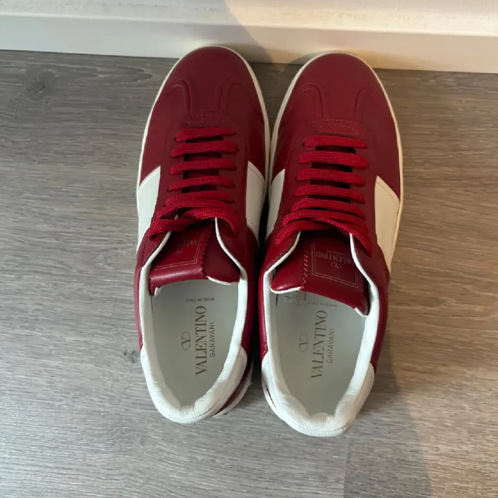 Säljer ett par snygga Valentino sneakers i rött och vitt skinn. De har coola nitar på hälen och en klassisk snörning framtill. Perfekta för att ge din outfit en lyxig touch. Skorna är i mycket bra skick och kommer med en grön dustbag. Passar perfekt för både vardag och fest!. Skor.