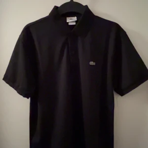 Svart pikétröja från Lacoste - Säljer en klassisk svart pikétröja från Lacoste i mycket bra skick. Den har korta ärmar och den ikoniska krokodilloggan på bröstet. Perfekt för både vardag och lite finare tillfällen. Passar till allt och är superbekväm!