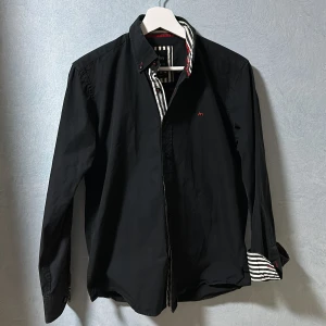 Svart Saint Martin Skjorta - Jag säljer en riktigt stilig svart sljorta från Saint Martin. Skjortan är svart med völdigt många riktigt snygga detaljer, som till exempel ärmarna och kragens svartvit randiga färg och Saint Martin loggan och knapparnas röda färg. Skick 9,5/10. M