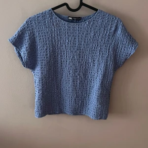 Blå topp från Zara - Säljer en snygg blå topp från Zara i ett texturerat material. Den har en rund hals och korta ärmar, perfekt för en avslappnad stil. Toppen är i bra skick och passar både till vardags och lite finare tillfällen.