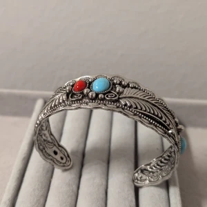 Silverfärgat armband med turkos och röd sten - Snyggt silverfärgat armband med detaljerade mönster och två stora stenar i turkos och rött. Armbandet har en vintagekänsla med sina intrikata snirkliga mönster och pärldetaljer.