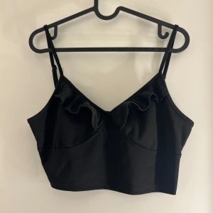 Svart croppad linnetopp med volang - Snygg svart croppad linnetopp med volangdetalj vid halsringningen. Perfekt för en trendig look.