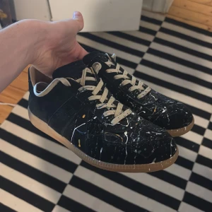 Maison margiela - Unika svarta sneakers från Maison Margiela med färgstänk i vitt, gult och blått. Skorna har beige snörning och en klassisk gummisula. Perfekta för dig som vill sticka ut med en konstnärlig touch.