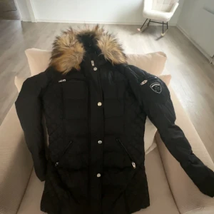 Svart dunjacka med päls från Rockandblue - Snygg svart dunjacka från Rockandblue med avtagbar päls på huvan. Jackan har dragkedja och knappar framtill samt praktiska fickor med dragkedja. Perfekt för kalla vinterdagar.