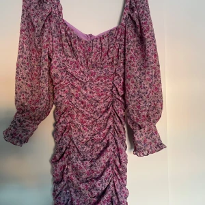 Blommig klänning från Zara - Säljer en vacker blommig klänning från Zara i rosa och lila toner. Klänningen har långa ärmar och en rynkad design som ger en fin siluett. Perfekt för en somrig look!
