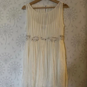 Vit ärmlös klänning med pärldetaljer - Elegant vit ärmlös klänning med vackra pärldetaljer runt midjan. Den har en luftig och flödande design som ger en romantisk känsla. Perfekt för speciella tillfällen.