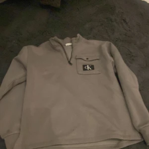 Grå tröja från Calvin Klein - Snygg grå tröja från Calvin Klein med dragkedja och en liten bröstficka med CK-logga. Perfekt för en avslappnad stil.
