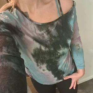 Flerfärgad offshoulder topp - Säljer en cool offshoulder topp med batikmönster i svart, blått och lila. Toppen har långa ärmar och en avslappnad passform, perfekt för en avslappnad stil.