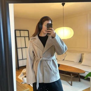 Helt ny jacka från Nakd - Snygg beige kappa med knappar och bälte i midjan. Perfekt för höst och vår. Den har en klassisk krage och långa ärmar, vilket ger en stilren look.