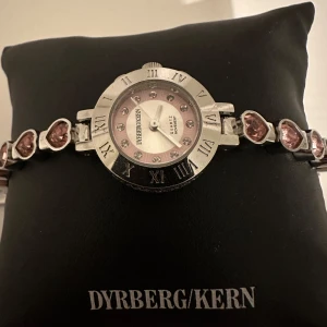 Silverfärgad klocka med hjärtan från Dyrberg/Kern - Elegant klocka från Dyrberg/Kern med silverfärgad boett och armband dekorerat med rosa hjärtan. Urtavlan har romerska siffror och glittriga stenar som timmarkeringar. Perfekt för att ge en romantisk touch till din stil. Behöver bytas batteri men annars felfri. Nypris 2000kr