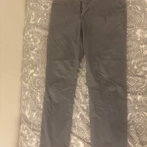 Grå chinos från Tommy Hilfiger - Snygga grå chinos från Tommy Hilfiger i slim fit. Byxorna har en klassisk design med knappar bak och bälteshällor. Perfekta för en stilren look.